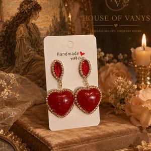 May include: A pair of gold-tone drop earrings on a white display card. Each earring features a small, oval-shaped red crystal stud connected to a larger, heart-shaped red pendant. Both the stud and the heart pendant are framed by tiny clear crystals.
