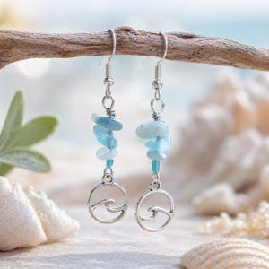 May include: A pair of silver dangle earrings. Each earring features a strand of light blue, irregular-shaped stones and a small, round, silver charm with a wave design. The earrings are suspended from silver ear wires and are displayed against a blurred beach background.