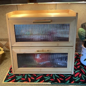 May include: A two-tiered bamboo bread box with glass paneled doors and gold-toned handles. The doors have a ribbed texture. The bread box sits on a black mat with a red and green chili pepper pattern. The top of the box has a small shelf.