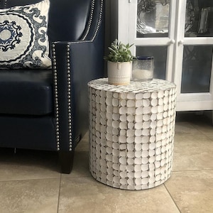 May include: A white, round side table with a textured, patterned surface. The table has a cylindrical shape and is placed next to a blue leather chair. A small potted plant and a glass are on top of the table.