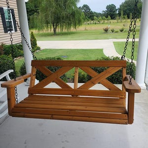 May include: A wooden porch swing with a natural finish, suspended by black chains. The swing features a slatted seat and a back with an X-shaped design. The armrests extend outward, offering a comfortable seating area. The swing is designed for outdoor use.
