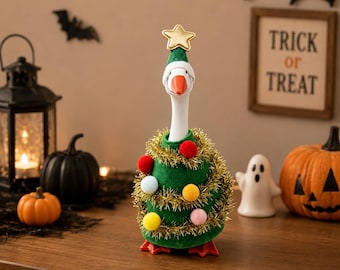 May include: A white goose figurine wearing a green felt cone shaped like a Christmas tree, decorated with gold tinsel and colourful pom-poms. The goose has an orange beak and a gold star on top of its head, standing on orange webbed feet.