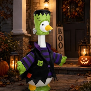 May include: A white goose figurine wearing a green Frankenstein-style hat with silver bolts, a purple and black striped long-sleeve shirt, and a black vest with a small green square patch featuring a smiling face and an orange square patch.