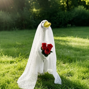 May include: A white goose figurine wearing a sheer white lace veil over its head and shoulders. The goose has a bright yellow beak and is holding a small bouquet of three vibrant red roses in its wings, standing upright in a grassy outdoor area.