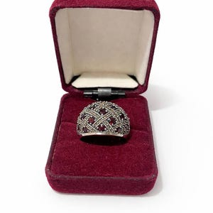May include: A silver ring with a domed top, set with small red stones and marcasite in a crisscross pattern. The ring is displayed in an open, burgundy velvet jewellery box with a cream-coloured interior.