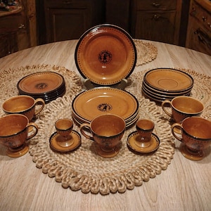 May include: A complete vintage dinnerware set in shades of brown and tan, featuring plates, saucers, cups, and egg cups. Each piece has a dark brown rim and a decorative emblem. The set is arranged on woven placemats.