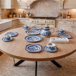 Könnte beinhalten: Ein runder Holztisch zeigt ein blau-weißes Teeservice, darunter eine Teekanne, Tassen, Untertassen und Teller. Das Set hat ein detailliertes Landschaftsdesign. Der Tisch steht in einer hellen Küche mit weißen Schränken und einem Kronleuchter.