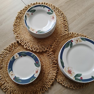 May include: A collection of white ceramic plates with a blue border, adorned with floral designs, are arranged on woven placemats. The plates feature a magnolia flower and green leaves. The placemats are made of natural fiber.