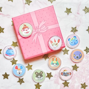 Puede incluir: Una colección de pines redondos con diseños de personajes de animales y criaturas, dispersos alrededor de una caja de regalo rosa con un lazo. El fondo es una superficie rosa claro con confeti dorado en forma de estrella.