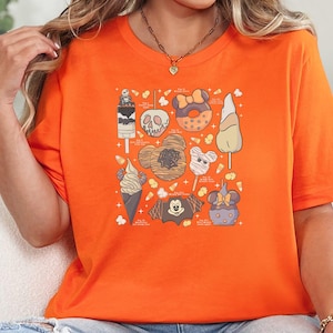 A woman wearing a bright orange tee with a Halloween-themed graphic collage featuring candy, skulls, pumpkins, ghost, and autumn icons in tan and gray.