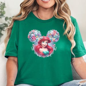 A woman wearing a kelly green tee with a graphic of a red-haired mermaid princess with mouse ear buns surrounded by colorful flowers.
