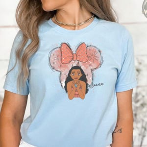 A woman wearing a light blue tee with an illustration of a girl with long black hair inside a mouse head with a red polka dot bow and the name "Moana" below.