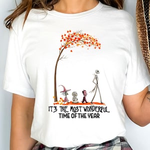 A woman wearing a white tee with an autumn tree and characters from The Nightmare Before Christmas with text reading "It's the most wonderful time of the year.