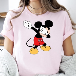 A woman wearing a light pink tee with a classic Mickey Mouse graphic showing the character with his arms raised wearing red shorts and yellow shoes.