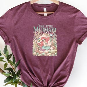 Heather maroon tee featuring "The Little Mermaid" title with Ariel swimming among coral and seaweed in vintage illustration style.