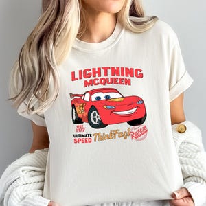 Woman wearing a cream tee with a red Lightning McQueen car graphic and text reading "Lightning McQueen est. 1977 Ultimate Speed ThinkFast Piston.