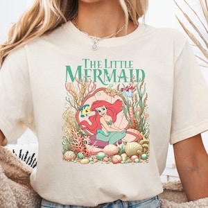 A woman wearing a cream Bella Canvas tee with "The Little Mermaid" text and a graphic of Ariel surrounded by colorful underwater coral and sea creatures.