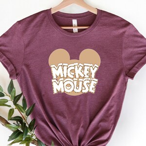 Heather maroon tee with tan Mickey Mouse ears and white outlined text reading "MICKEY MOUSE" across the chest.