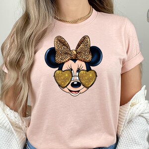 A woman wearing a light pink t-shirt featuring Minnie Mouse with gold glitter heart-shaped sunglasses and a leopard print bow, styled with denim shorts and a white knit cardigan.