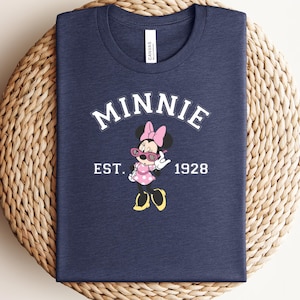 Flat lay of a navy blue t-shirt with Minnie Mouse graphic wearing pink sunglasses, bold white arched text reading "MINNIE" and "EST. 1928" on the chest, displayed folded on a wicker surface.