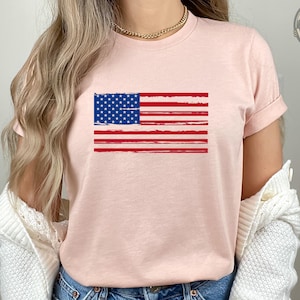 May include: A light pink t-shirt featuring a distressed American flag design in red, white, and blue. The shirt has short sleeves and a crew neck. The model is wearing blue denim shorts and a white cardigan.