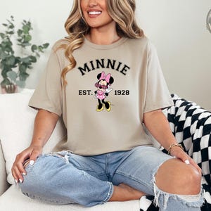 A woman wearing a tan t-shirt with "MINNIE EST. 1928" in black collegiate text and a Minnie Mouse graphic wearing pink sunglasses and a pink polka dot dress.
