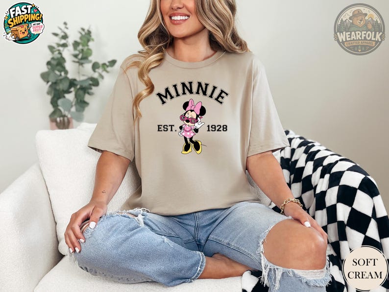 A woman wearing a tan t-shirt with "MINNIE EST. 1928" in black collegiate text and a Minnie Mouse graphic wearing pink sunglasses and a pink polka dot dress.