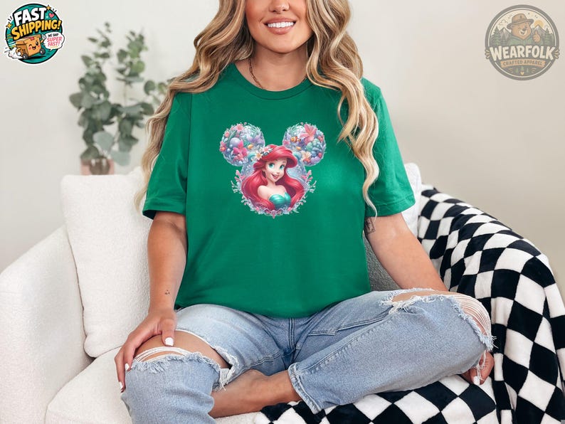 A woman wearing a kelly green tee with a graphic of a red-haired mermaid princess with mouse ear buns surrounded by colorful flowers.