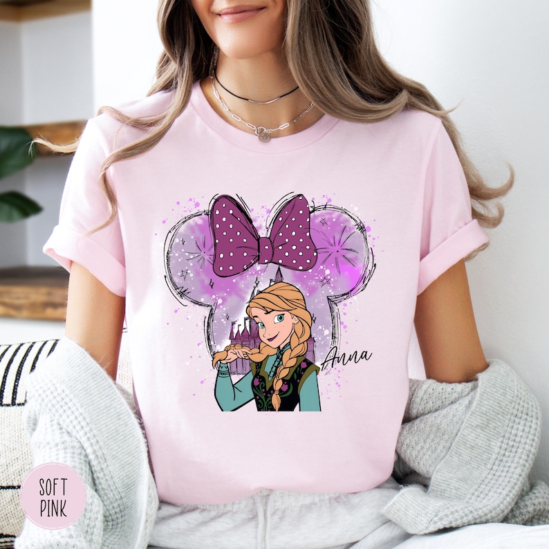 A woman wearing a soft pink tee with Anna from Frozen illustrated under a large purple polka-dot bow with sparkle accents and "Anna" text.