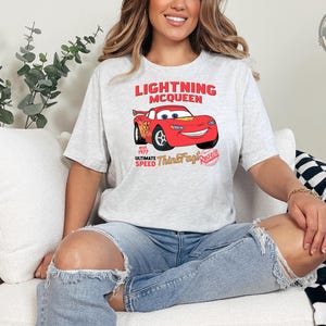 K&ouml;nnte beinhalten: Hellgraues T-Shirt mit Lightning McQueen aus dem Film Cars. Das Design zeigt den Schriftzug "Lightning McQueen" &uuml;ber einem roten Cartoon-Auto, darunter "est. 1977 Ultimate Speed Think Fast! Race". Das T-Shirt wird von einer Person getragen.