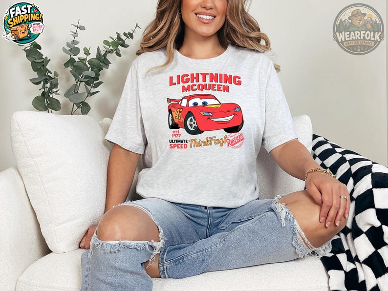 K&ouml;nnte beinhalten: Hellgraues T-Shirt mit Lightning McQueen aus dem Film Cars. Das Design zeigt den Schriftzug "Lightning McQueen" &uuml;ber einem roten Cartoon-Auto, darunter "est. 1977 Ultimate Speed Think Fast! Race". Das T-Shirt wird von einer Person getragen.