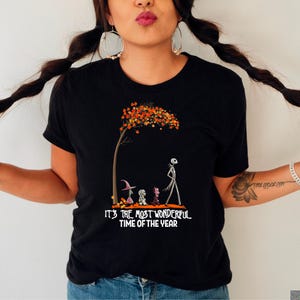 A woman wearing a black tee with a skeletal figure under an orange tree and characters below text reading "It's the most wonderful time of the year".