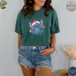 A woman wearing a dark green Bella Canvas tee with a blue alien character wearing a Santa hat and holding a candy cane surrounded by snowflakes.