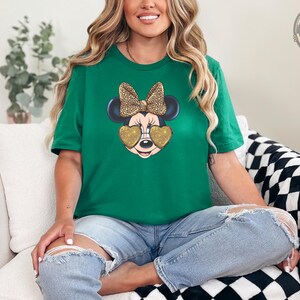 A woman wearing a kelly green t-shirt featuring Minnie Mouse with gold glitter heart-shaped sunglasses and a leopard print bow, sitting cross-legged on a white chair in ripped jeans.