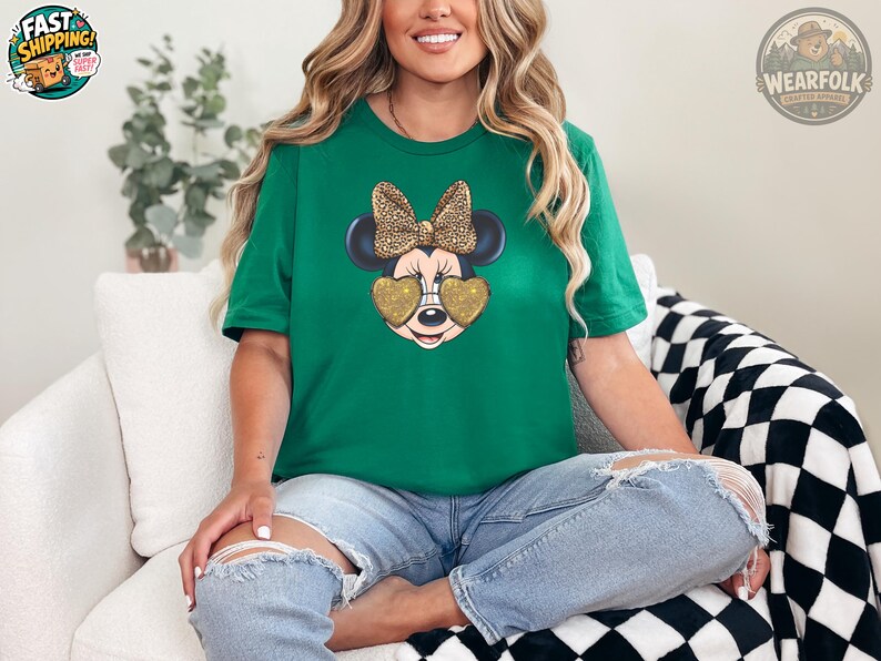 A woman wearing a kelly green t-shirt featuring Minnie Mouse with gold glitter heart-shaped sunglasses and a leopard print bow, sitting cross-legged on a white chair in ripped jeans.