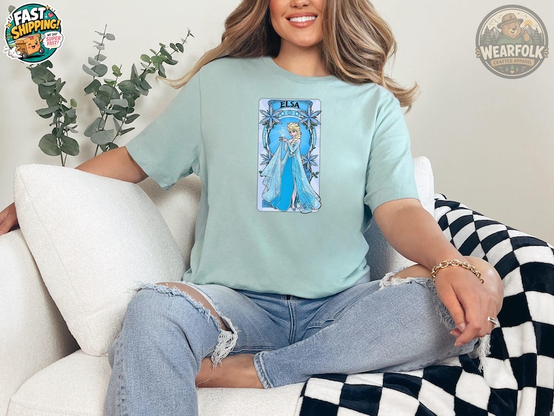 A woman wearing a light blue tee with an illustration of Elsa in a blue gown surrounded by snowflakes and the text "ELSA" at the top.