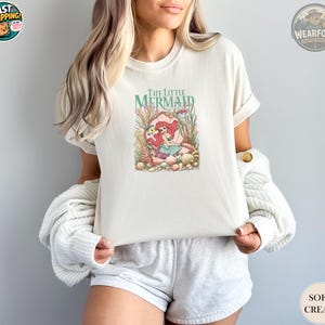 A woman wearing a soft cream Bella Canvas tee with a colorful illustration of Ariel from The Little Mermaid surrounded by underwater plants and coral.