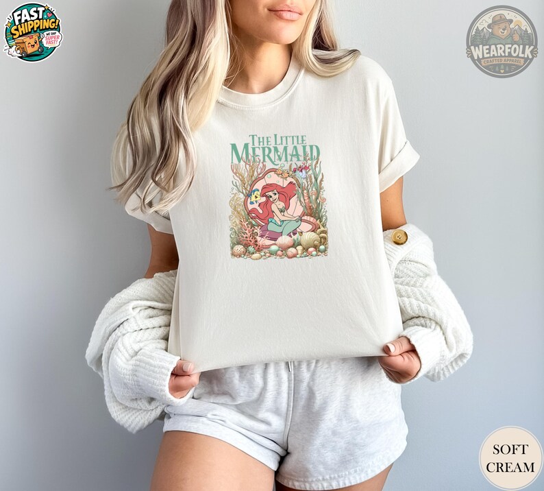 A woman wearing a soft cream Bella Canvas tee with a colorful illustration of Ariel from The Little Mermaid surrounded by underwater plants and coral.