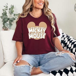 A woman wearing a burgundy tee with a tan Mickey Mouse head graphic and white "Mickey Mouse" text across the chest.