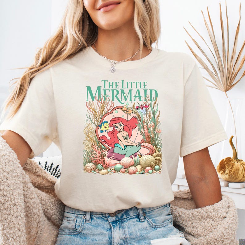 A woman wearing a cream Bella Canvas tee with "The Little Mermaid" text and a graphic of Ariel surrounded by colorful underwater coral and sea creatures.