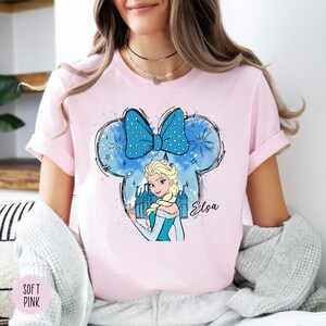 A woman wearing a soft pink tee with Elsa from Frozen inside a Mickey Mouse head shape with a blue bow and castle graphic.