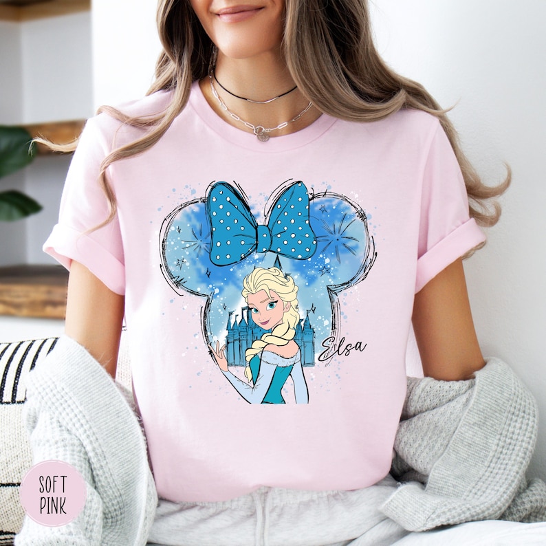 A woman wearing a soft pink tee with Elsa from Frozen inside a Mickey Mouse head shape with a blue bow and castle graphic.