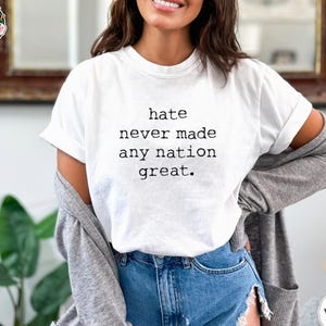 May include: A white short-sleeved t-shirt featuring the black printed text "hate never made any nation great." The shirt is worn with a gray long-sleeved cardigan and blue denim shorts, creating a casual layered look with a clear message.