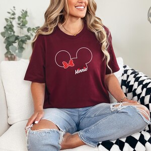 A woman wearing a burgundy tee with a Minnie Mouse head graphic featuring a red polka-dot bow and the word "Minnie" in white text.