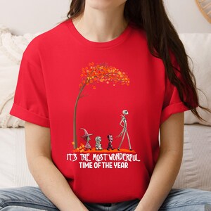 A woman wearing a red tee with a skeletal character and Halloween figures beneath a fall tree, text reading "It's the most wonderful time of the year".