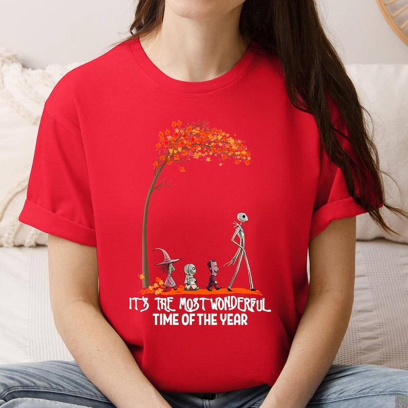 A woman wearing a red tee with a skeletal character and Halloween figures beneath a fall tree, text reading "It's the most wonderful time of the year".