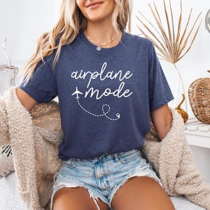 A woman wearing a heather navy t-shirt that says "airplane mode" in white cursive script with a small airplane graphic and dashed flight path design.