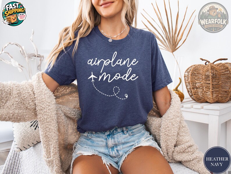 A woman wearing a heather navy t-shirt that says "airplane mode" in white cursive script with a small airplane graphic and dashed flight path design.