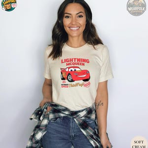 A woman wearing a cream tee with a red Lightning McQueen race car graphic and text that says "Lightning McQueen Ultimate Speed Thundering Racing.