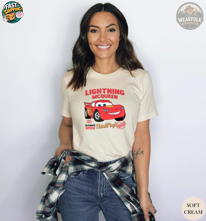 A woman wearing a cream tee with a red Lightning McQueen race car graphic and text that says "Lightning McQueen Ultimate Speed Thundering Racing.